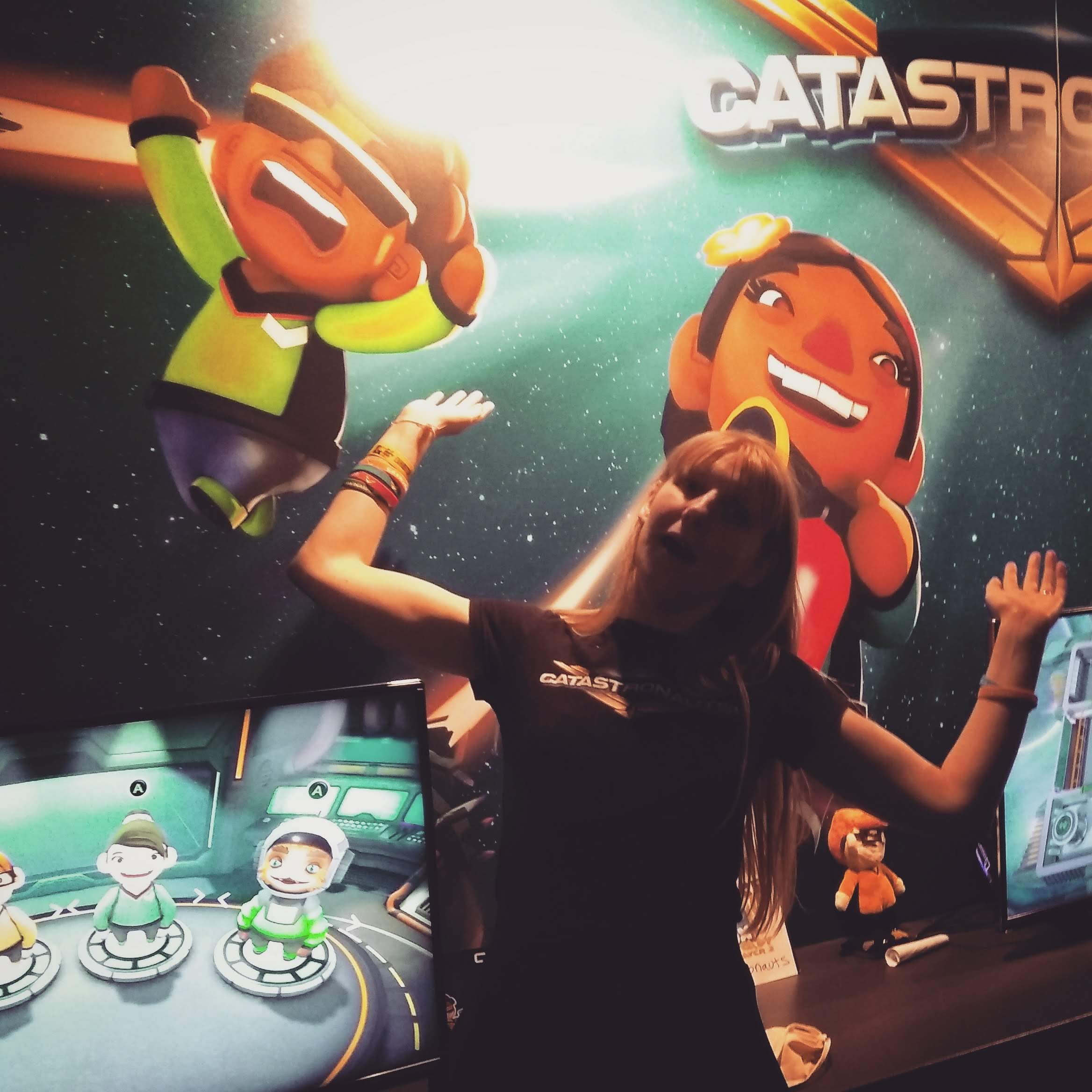 Kim posing, screaming with her arms up in the air, matching the Catastronaut on the poster behind her