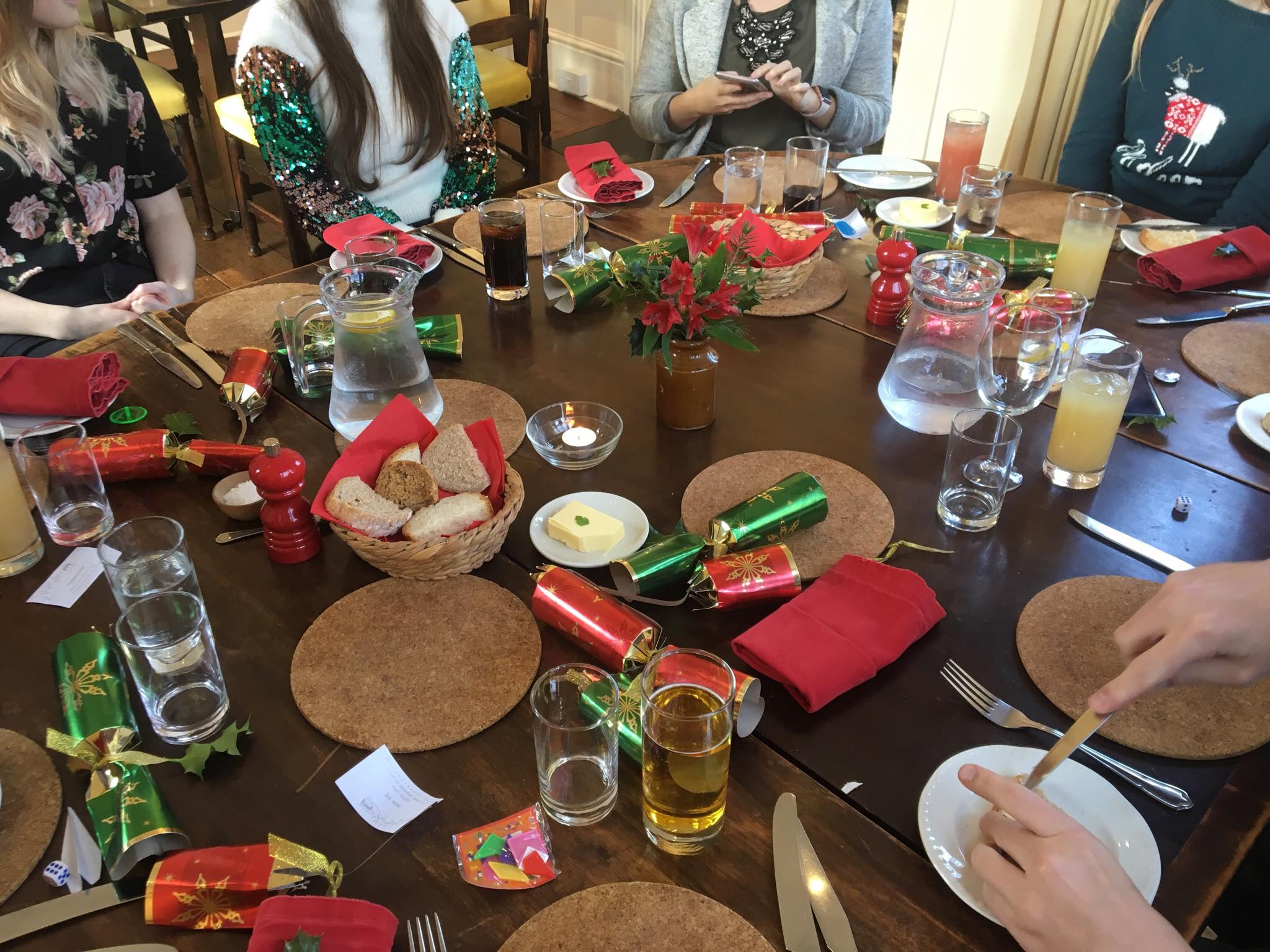 A round wooden table covered in cork placemats, glasses of various drinks and red and green napkins, Christmas crackers and flowers. People are seated around the edge wearing Christmassy clothes
