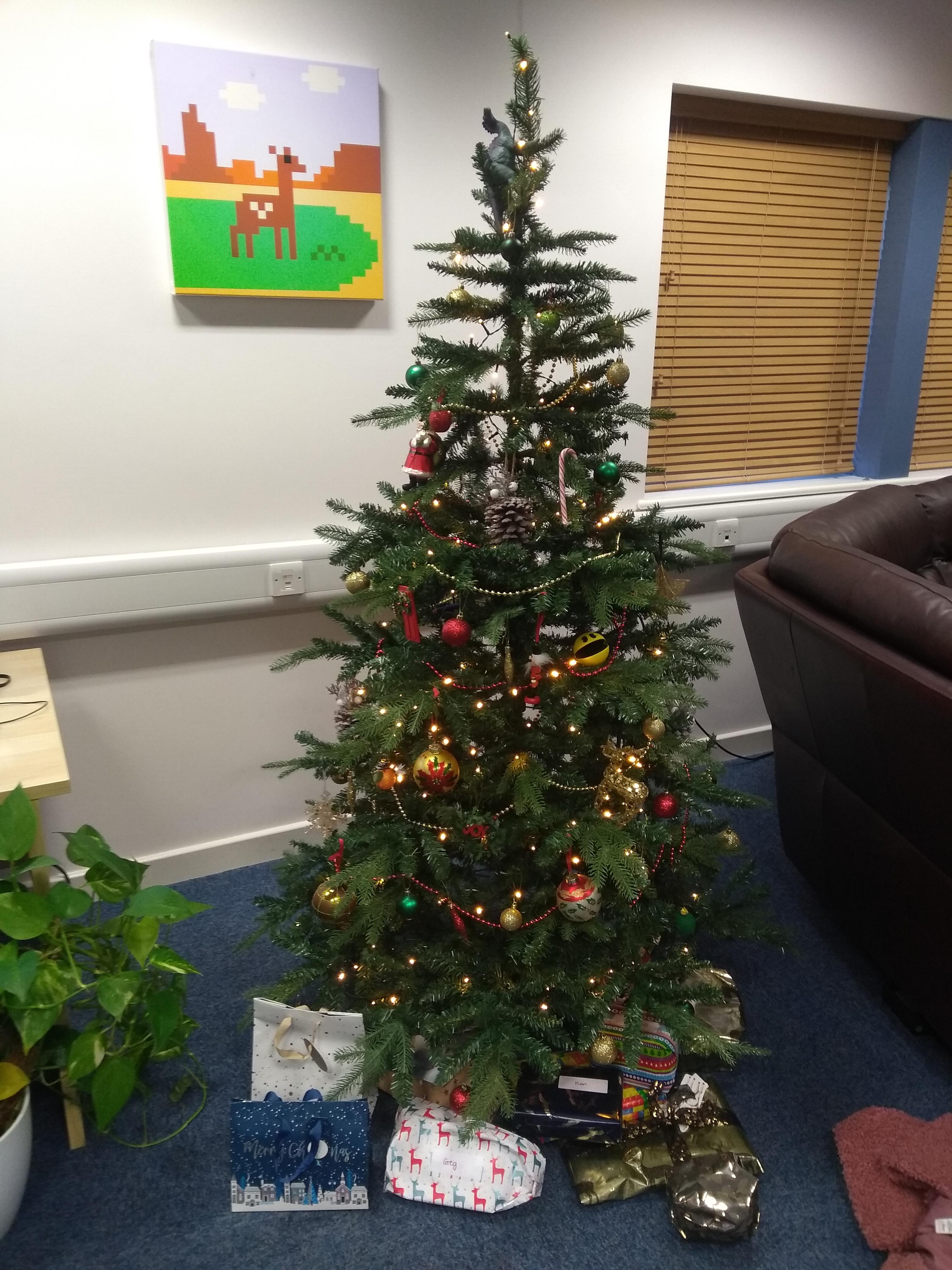 An artificial tree decorated with red and gold decorations and fairy lights. There are various wrapped gifts underneath, a dinosaur near the top of the tree and a Pacman hidden in the branches. It's placed in an office with white walls and a blue carpet.