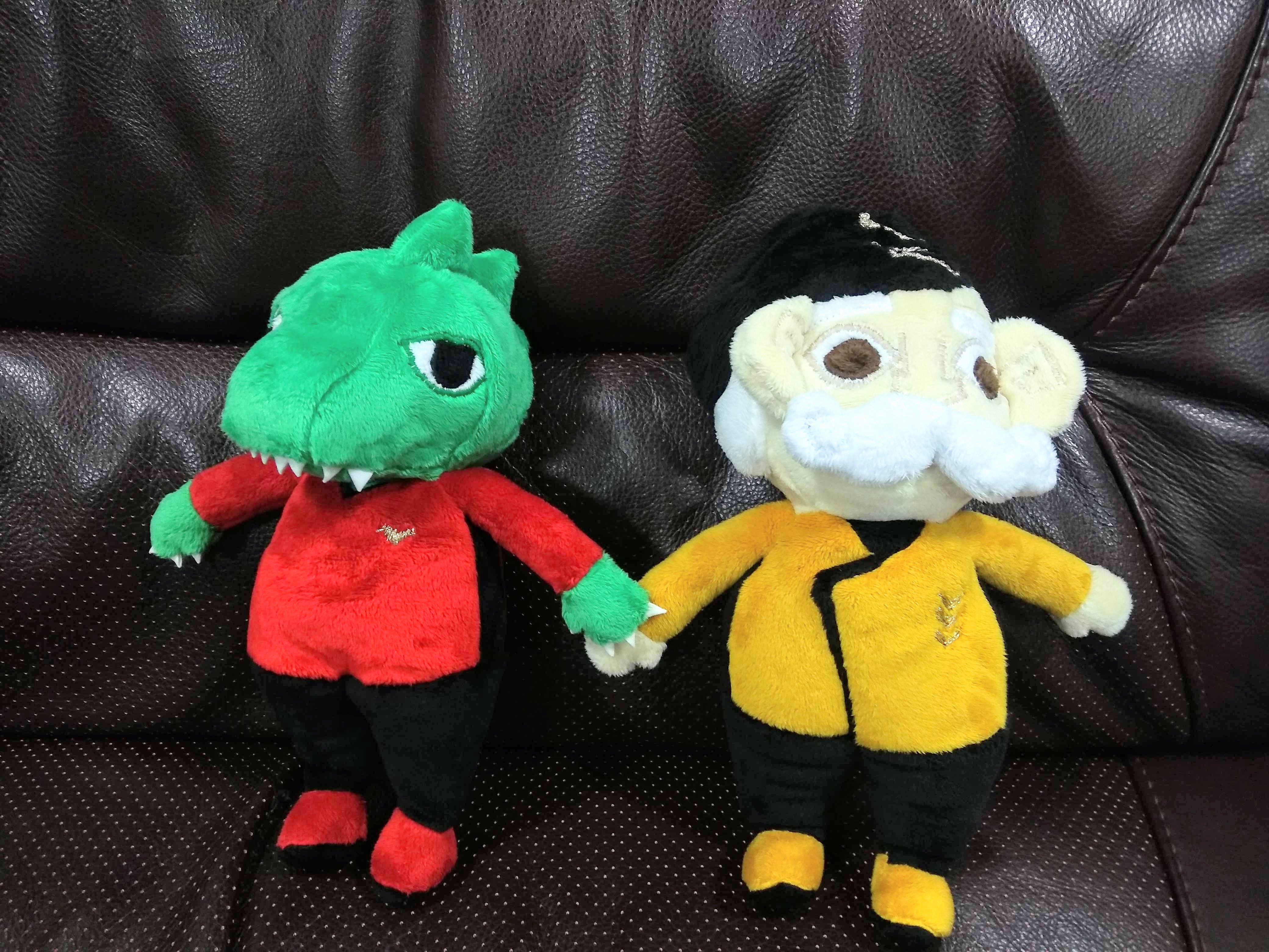 Two plushies on a leather sofa, both in 'Catastronauts' uniforms. In yellow and black is Sarge with a black hat, pale skin and white hair and large moustache. In red is a dinosaur-like green lizard with sharp teeth and ridges on his head.