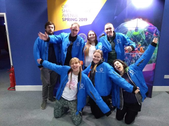 The Inertia team in blue Crystal Maze bomber jackets, posing with excited expressions (except two men who look confused) and arms out, in front of a picture of the Crystal Dome.