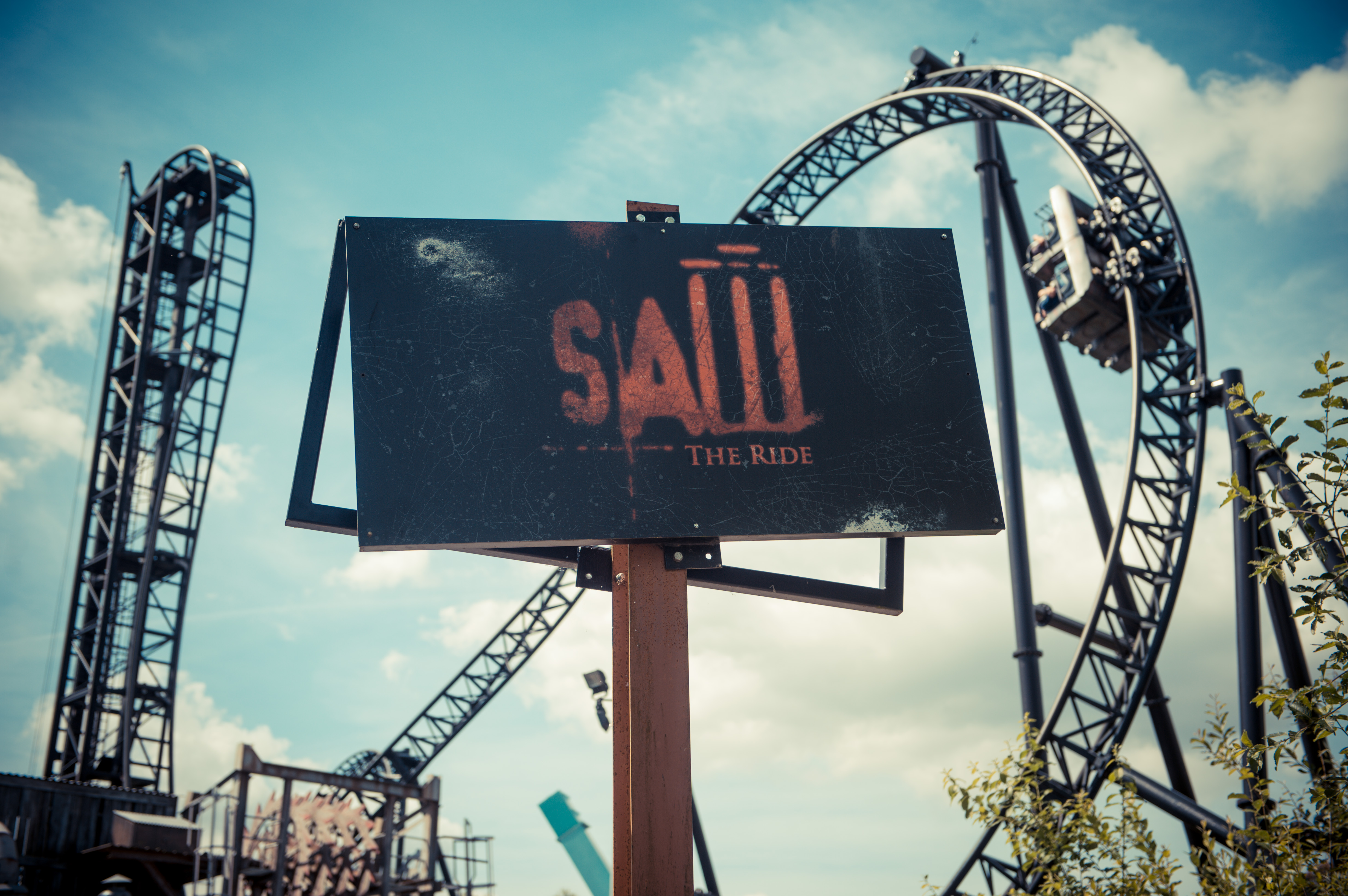 Roller coaster in brown metal with a loop on the right which is currently being climbed by a small cart and a tall straight up - straight down drop arch on the left. In the foreground is a sign reading SAW - The Ride.