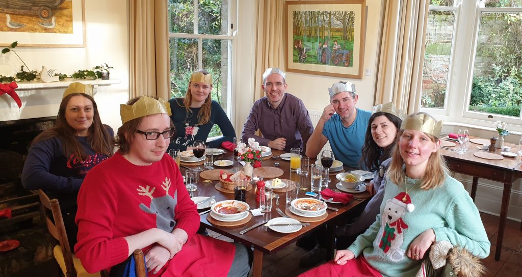 Group of people around a dining table full of tablewear, everyone is wearing a paper crown and several have on Christmas jumpers. They're in an old building with sash windows