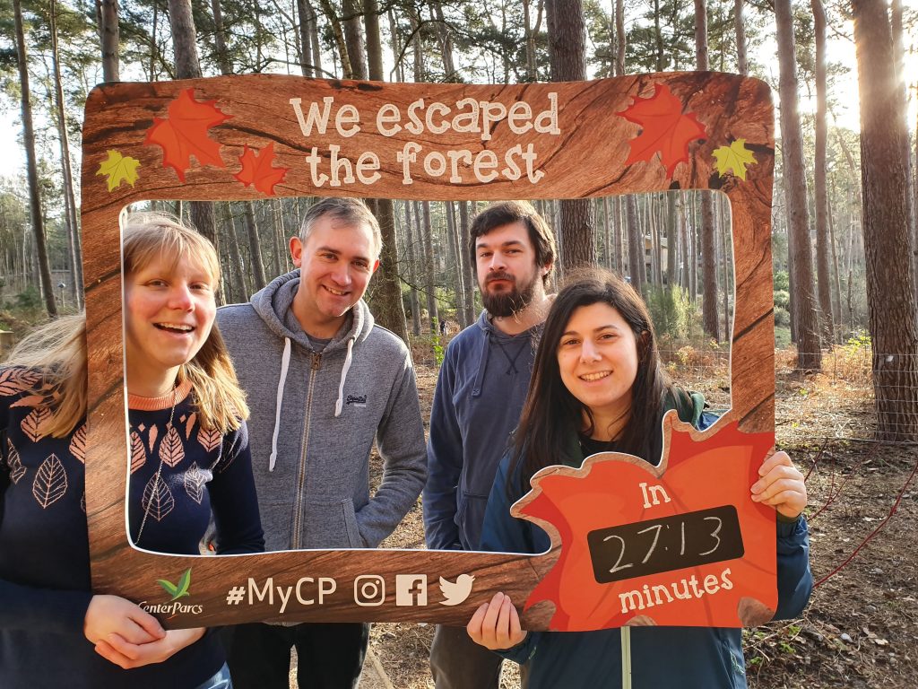 Four people smiling inside a large frame they are holding. It's decorated in a forest theme with "We escaped the forest in 27:13 minutes" written on it and the Center Parcs logo.