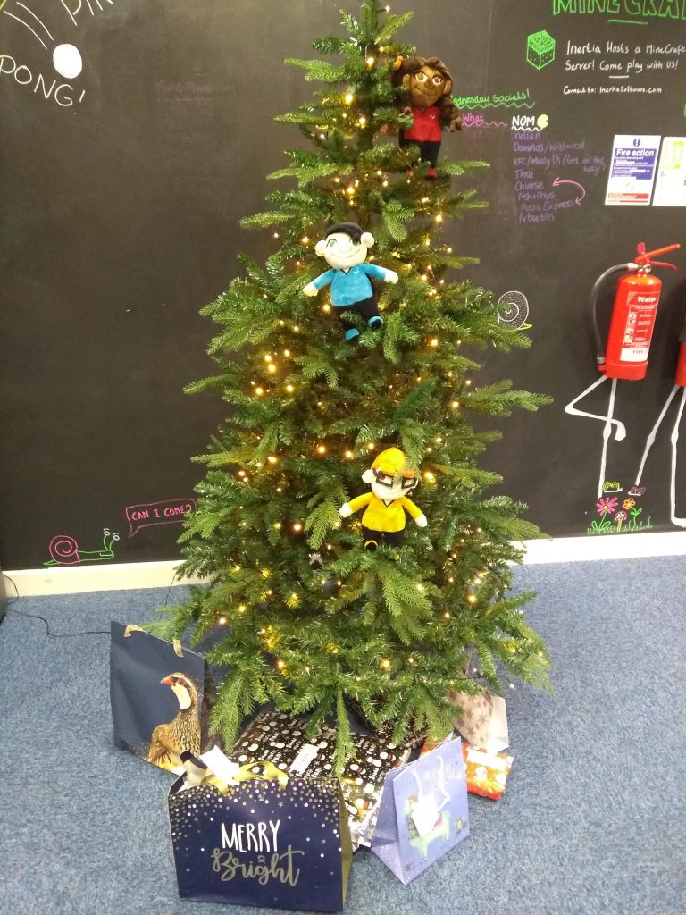 A Christmas tree with lights and three Catastronauts plushies on it, and wrapped gifts underneath it.